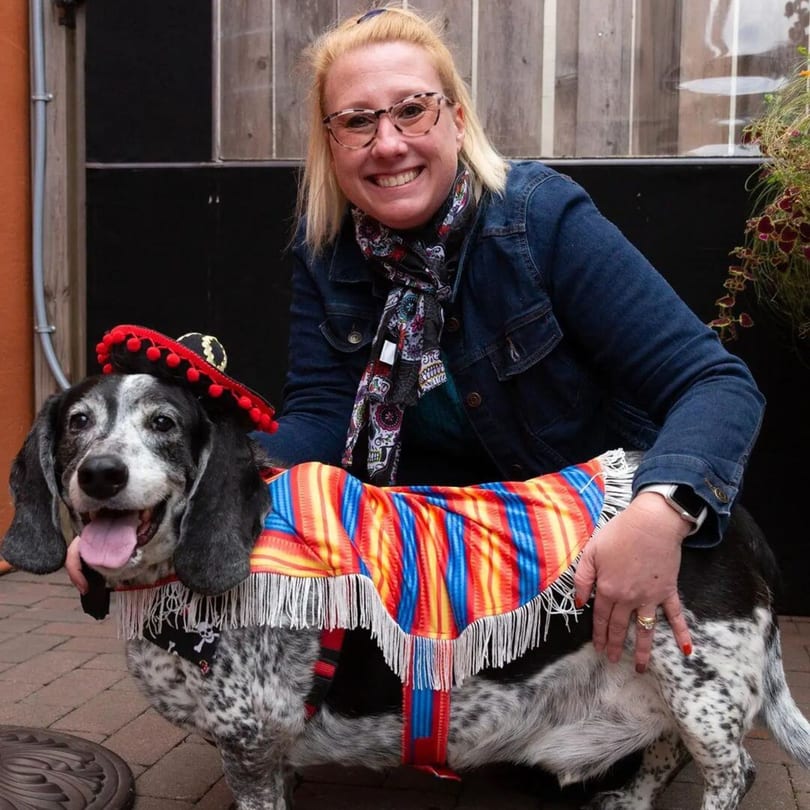Smiling woman petting a dog dressed in a colorful Mexican serape and sombrero.