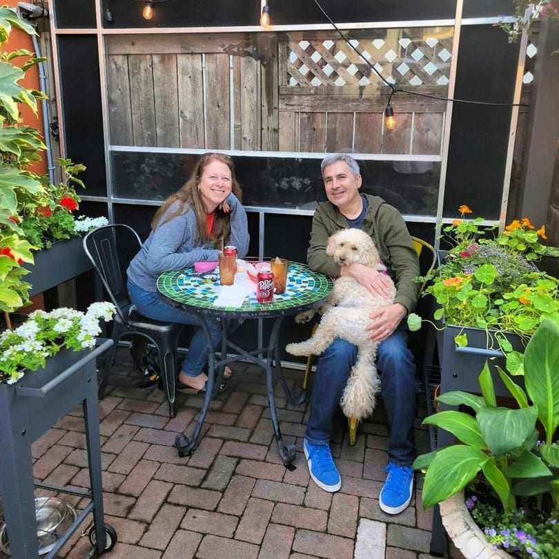 A couple and their dog sit at a mosaic table on a brick patio surrounded by plants at Tinga Fresh Mexican.