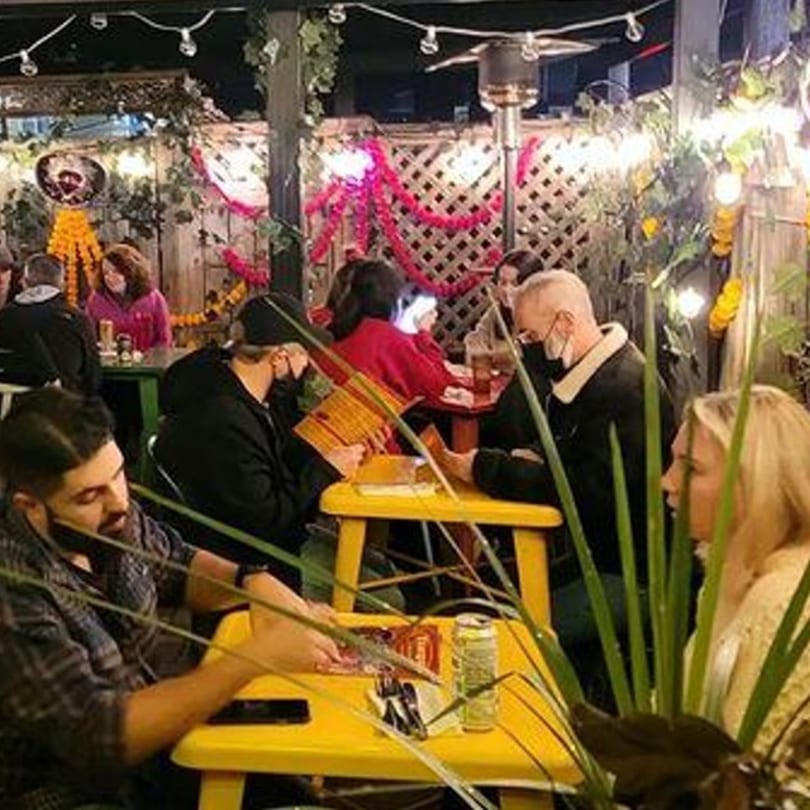 Diners seated at yellow tables on Tinga Fresh Mexican's outdoor patio, adorned with string lights and festive decor.