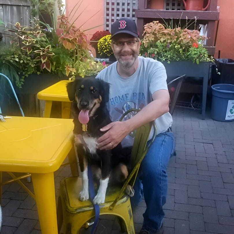 Man in a baseball cap and glasses petting a black and tan dog on a yellow chair outdoors.